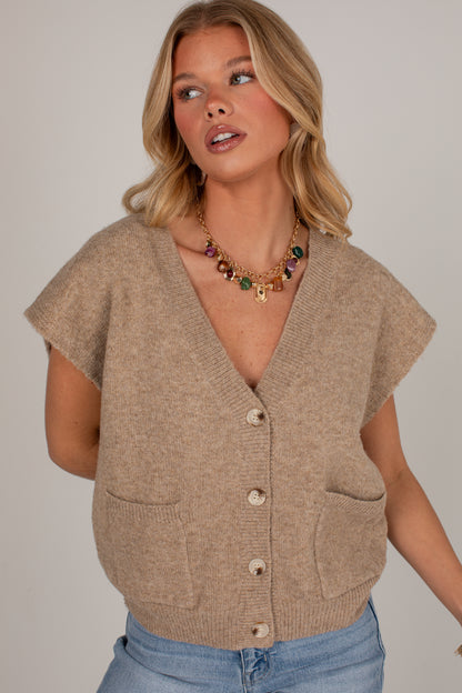A woman wearing a beige knit cardigan vest with short sleeves, a V-neck, front buttons, and patch pockets, paired with blue jeans against a light gray studio background.
