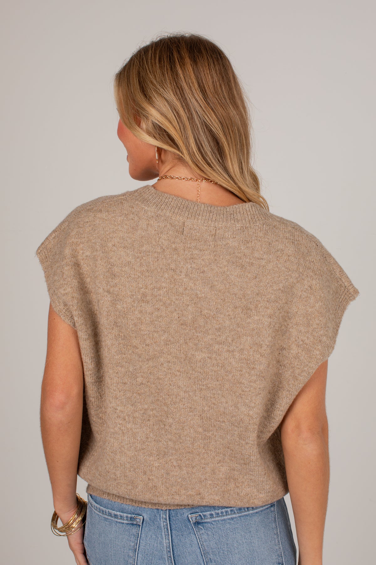 A woman viewed from the back wearing a light brown knit sweater with wide cap sleeves and light blue jeans, set against a plain light gray studio background.