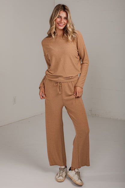 A woman modeling a matching tan knit lounge set consisting of a long-sleeve top and wide-leg drawstring pants, paired with beige sneakers, in a bright studio setting.