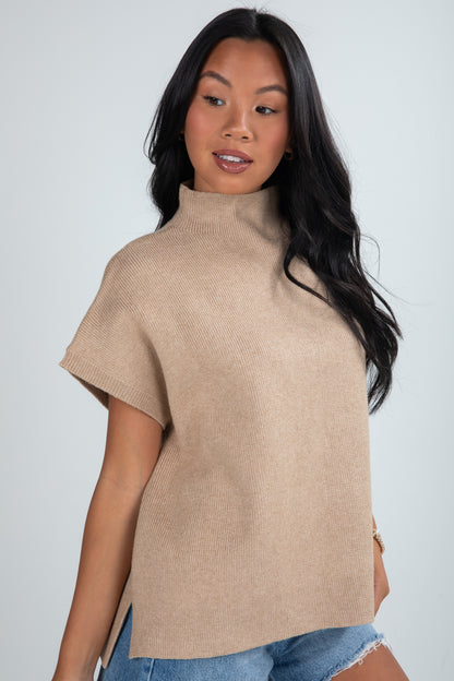 Woman wearing a beige turtleneck sweater over a blue top against a plain background