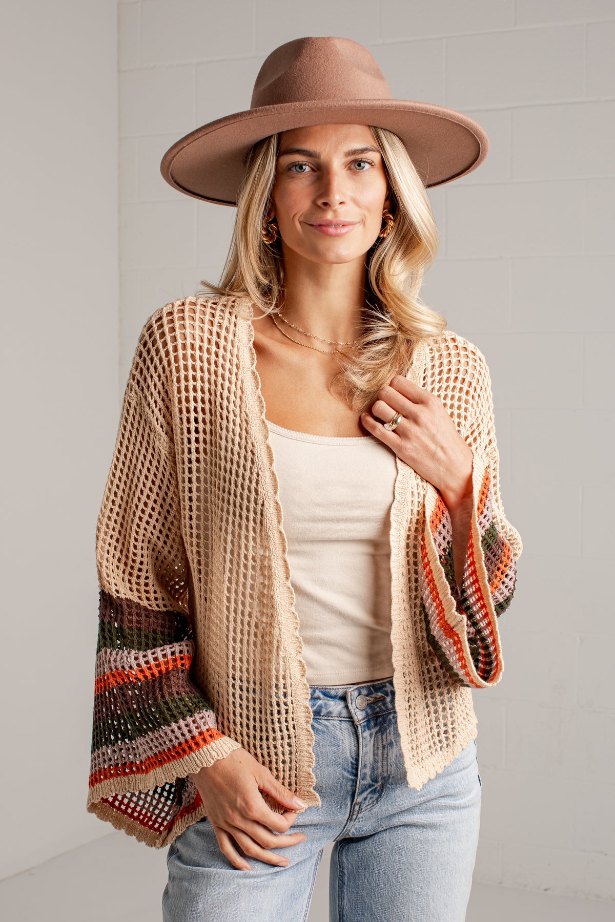 A woman modeling a beige open-knit cardigan with colorful striped bell sleeves, styled with a brown wide-brim hat, a cream tank top, and light-wash blue jeans against a white brick wall.