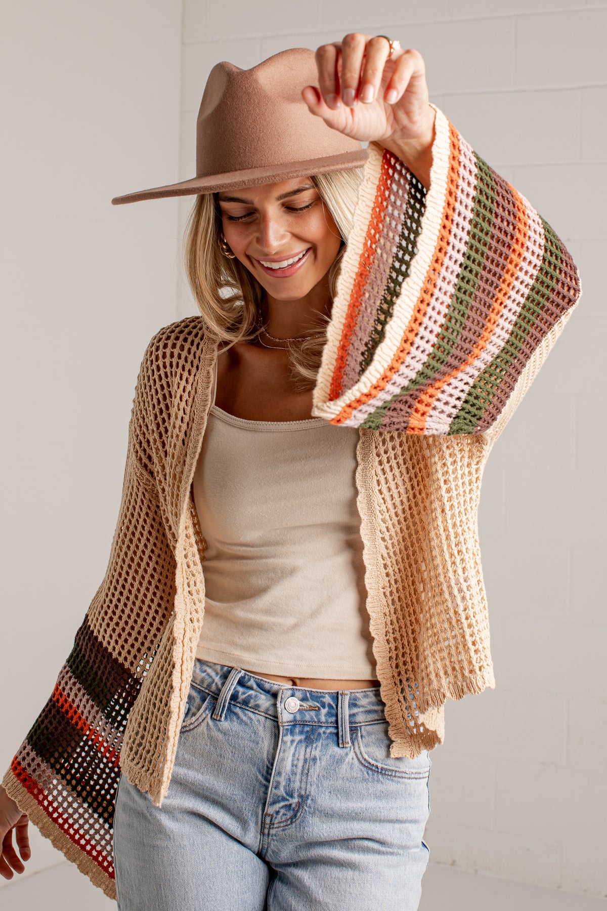 A woman wearing a chunky knit open-front cardigan with striped knit cuffs and hem, a beige hat, and light-wash jeans, posing against a white wall.