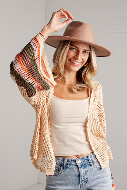 A woman wearing a cream open-knit cropped cardigan with colorful striped knit sleeves, a beige wide-brim fedora hat, and a cream tank top paired with light-wash blue jeans, posing against a white paneled wall.