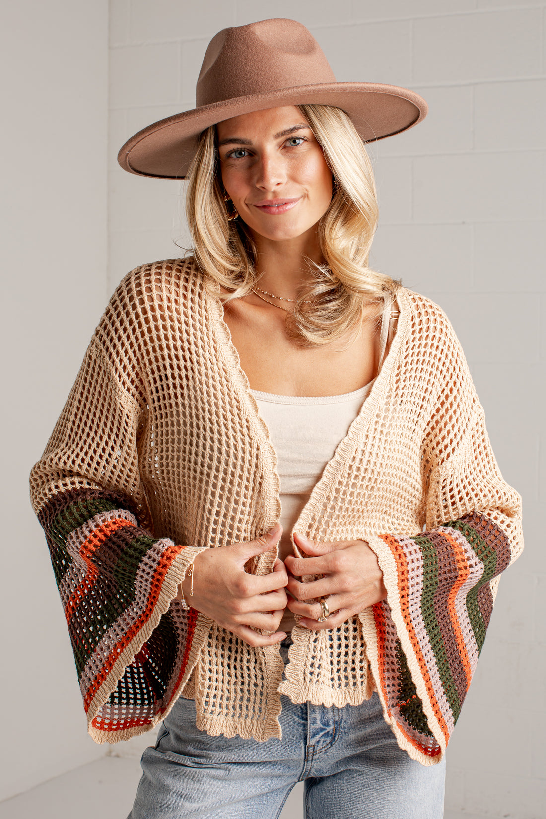 A woman wearing a beige open-knit cropped cardigan with wide bell sleeves featuring multicolor striped cuffs, a brown wide-brim hat, and light blue jeans, standing against a white brick wall.