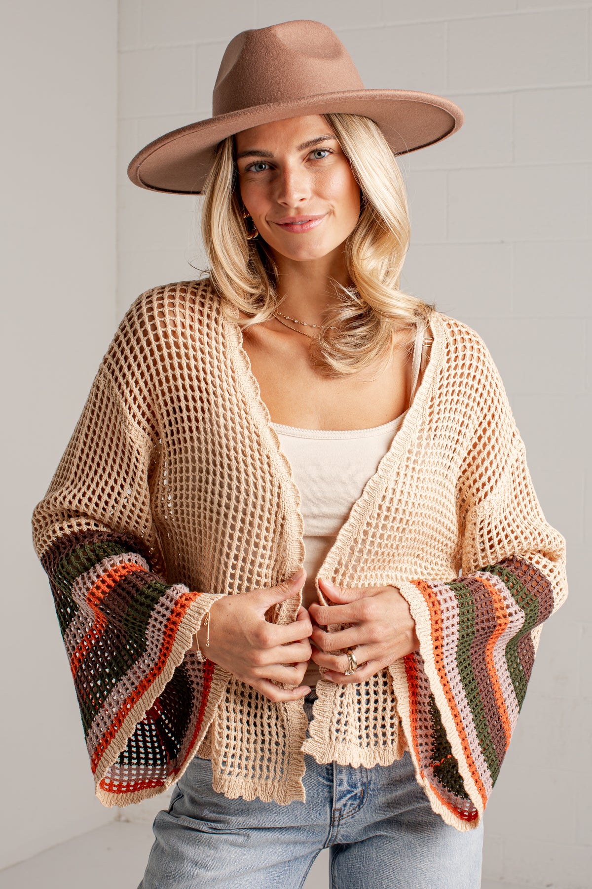 A woman wearing a beige open-knit cropped cardigan with wide bell sleeves featuring multicolor striped cuffs, a brown wide-brim hat, and light blue jeans, standing against a white brick wall.