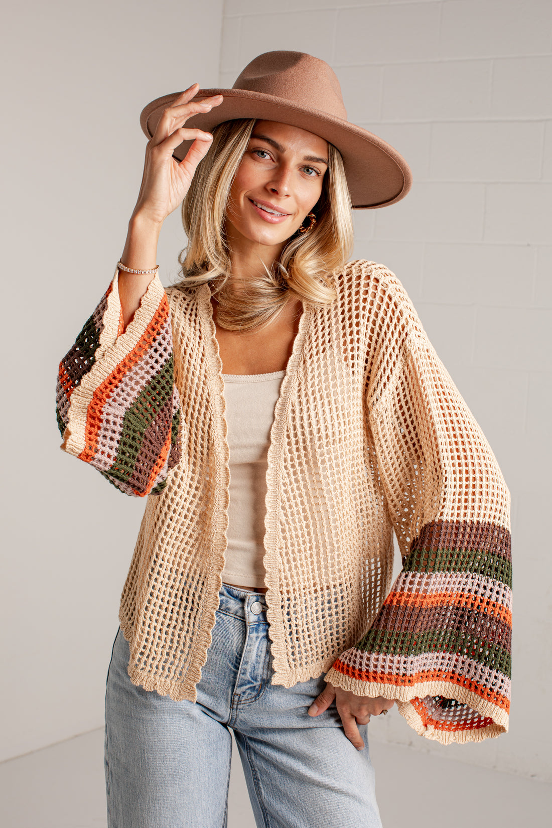 A woman wearing a beige open-knit cropped cardigan with colorful striped bell sleeves, styled with a brown wide-brim hat, a beige tank top, and light-wash blue jeans against a white brick wall.