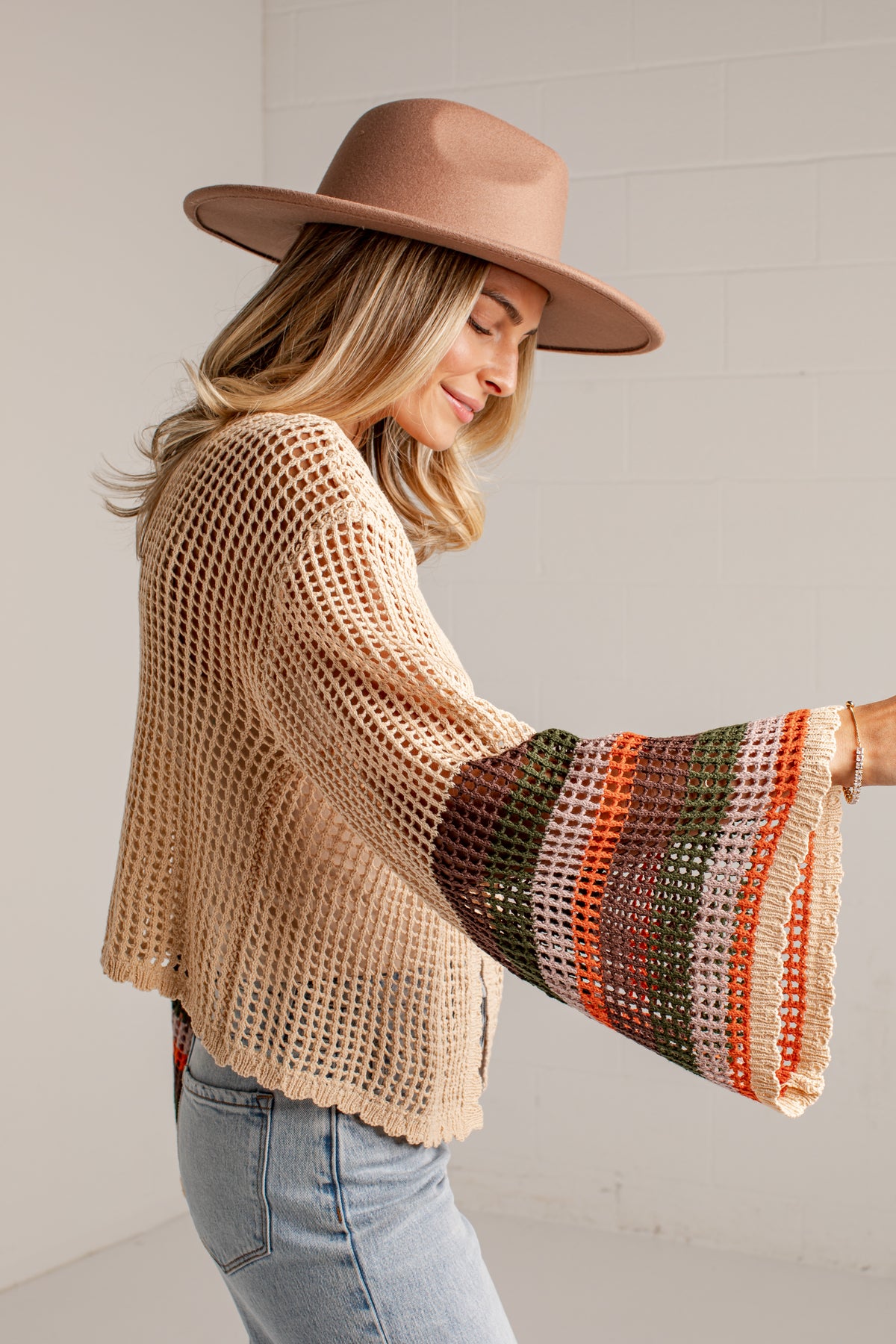 A woman wearing a beige open-knit cropped cardigan with wide bell sleeves featuring striped knit patterns in orange, green, and cream, paired with a brown wide-brim hat and light-wash jeans.