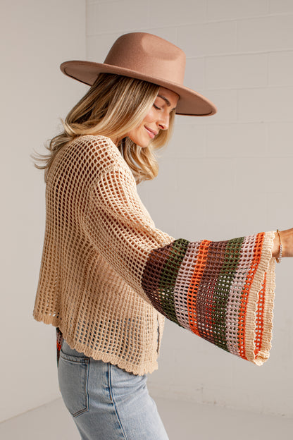 A woman wearing a beige open-knit cropped cardigan with wide bell sleeves featuring striped knit patterns in orange, green, and cream, paired with a brown wide-brim hat and light-wash jeans.