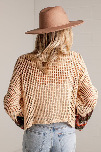 A woman shown from behind wearing a beige open-knit cropped sweater, a tan wide-brim hat, and light-wash blue jeans against a light gray wall.