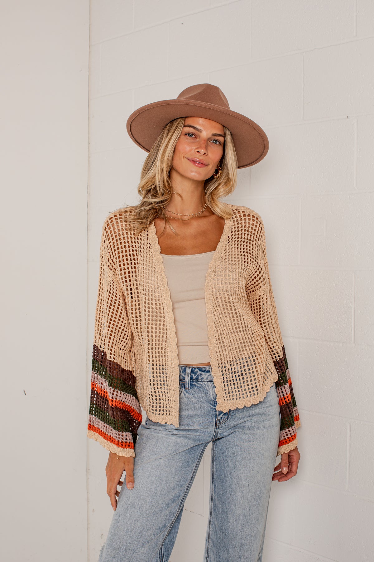 A woman wearing a beige open-knit cardigan with colorful striped cuffs, a brown wide-brim hat, a beige tank top, and light blue jeans, standing against a white brick wall.