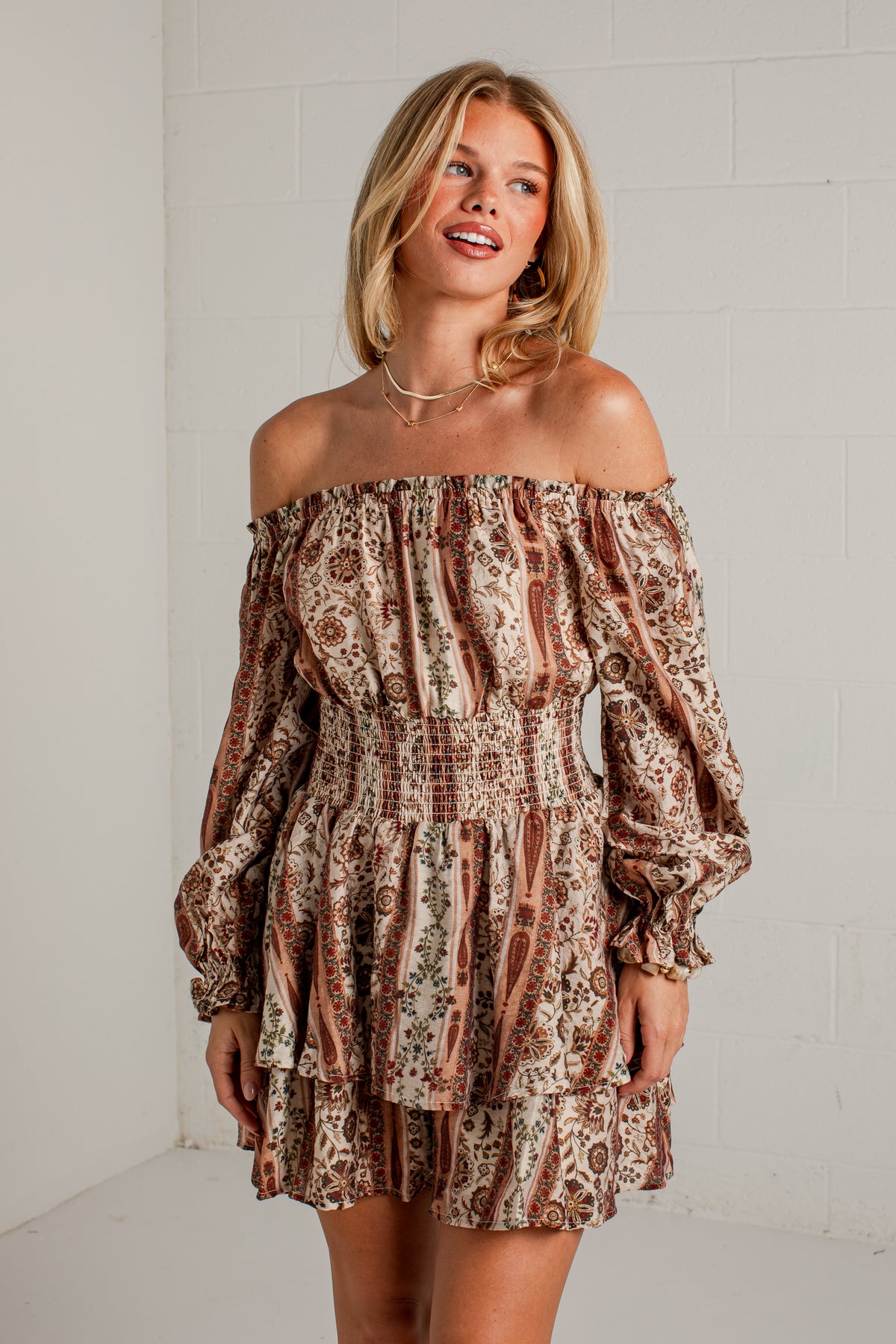 A woman modeling an off-the-shoulder mini dress with a brown, beige, and black paisley pattern, standing against a white brick wall.