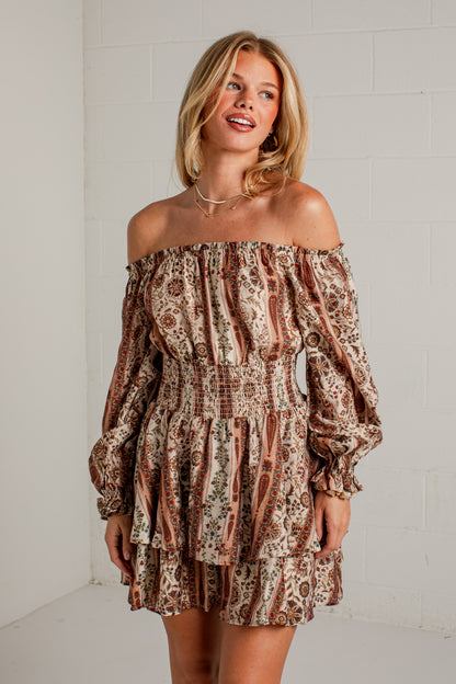 A woman modeling an off-the-shoulder mini dress with a brown, beige, and black paisley pattern, standing against a white brick wall.