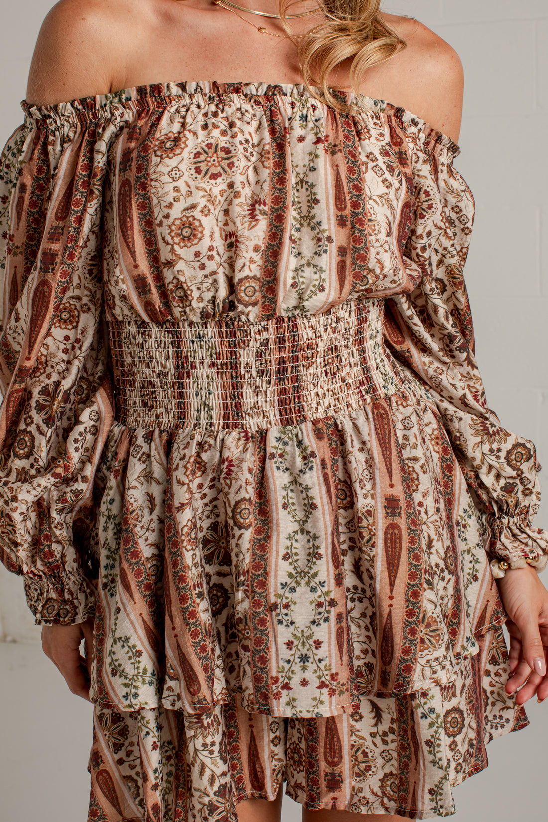 A woman modeling an off-the-shoulder mini dress with a brown and beige paisley pattern, paired with beige ankle boots, standing against a white brick wall.