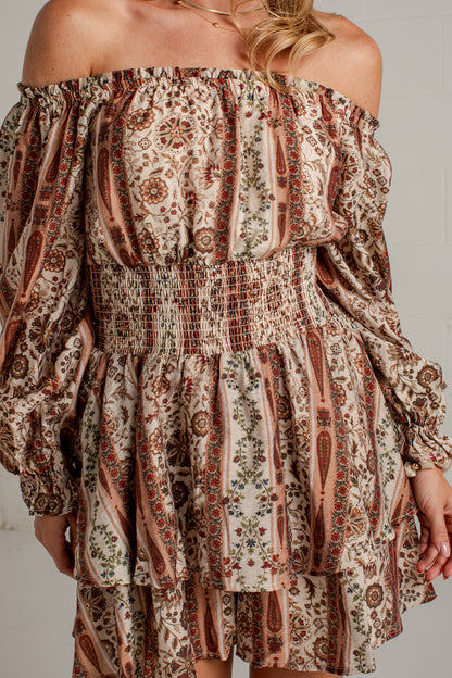 A woman modeling an off-the-shoulder mini dress with a brown and beige paisley pattern, paired with beige ankle boots, standing against a white brick wall.