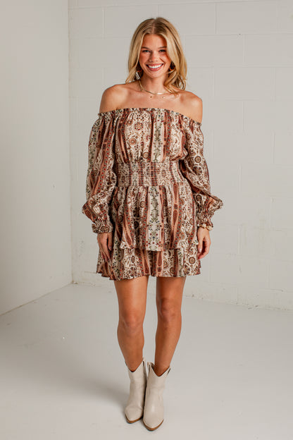 A woman modeling an off-the-shoulder mini dress with a brown and beige paisley pattern, paired with beige ankle boots, standing against a white brick wall.