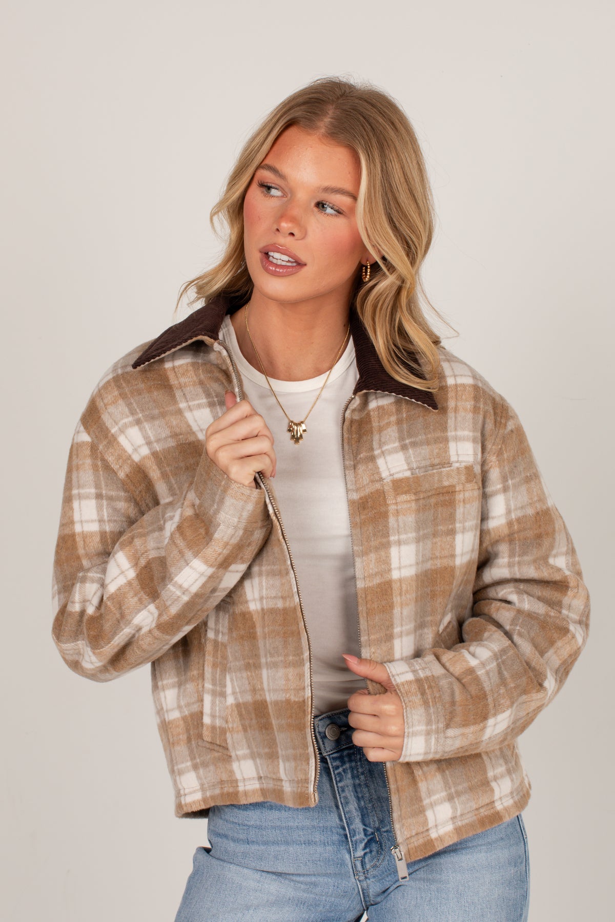 A woman modeling a cropped, zip-front plaid jacket with a brown collar, worn over a white t-shirt and blue jeans, against a light gray studio background.
