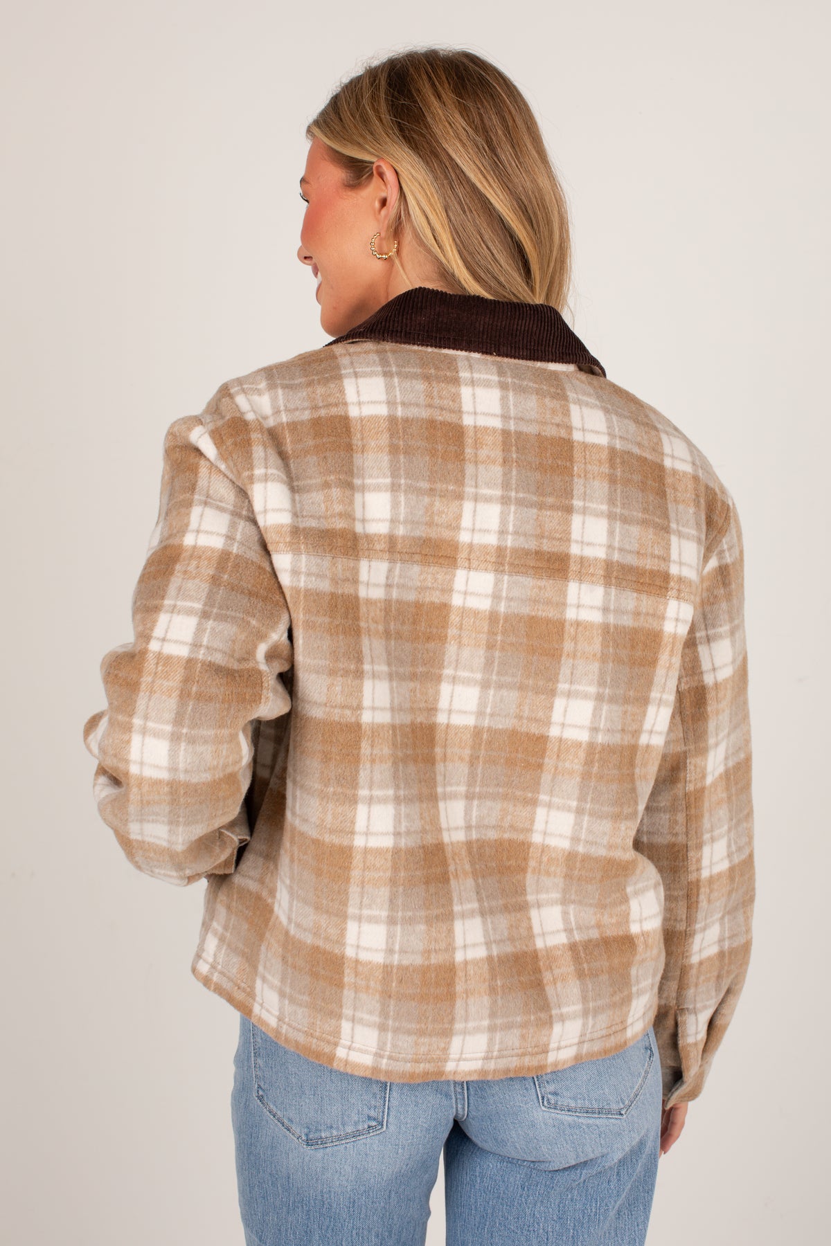A woman shown from the back wearing a tan and cream plaid flannel shirt jacket with a dark brown corduroy collar, paired with blue jeans, against a plain light gray background.