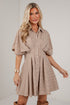 A woman modeling a beige and white striped short-sleeve button-up dress with a flared skirt, standing against a white paneled wall.