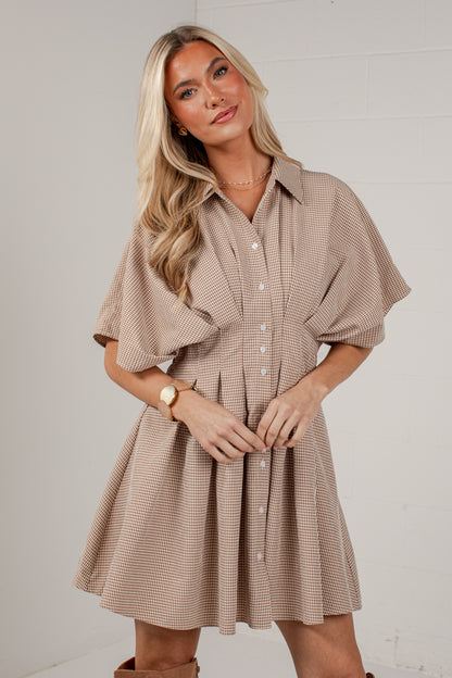A woman modeling a beige and brown striped short-sleeve button-up dress with a flared skirt, standing against a light gray brick wall.