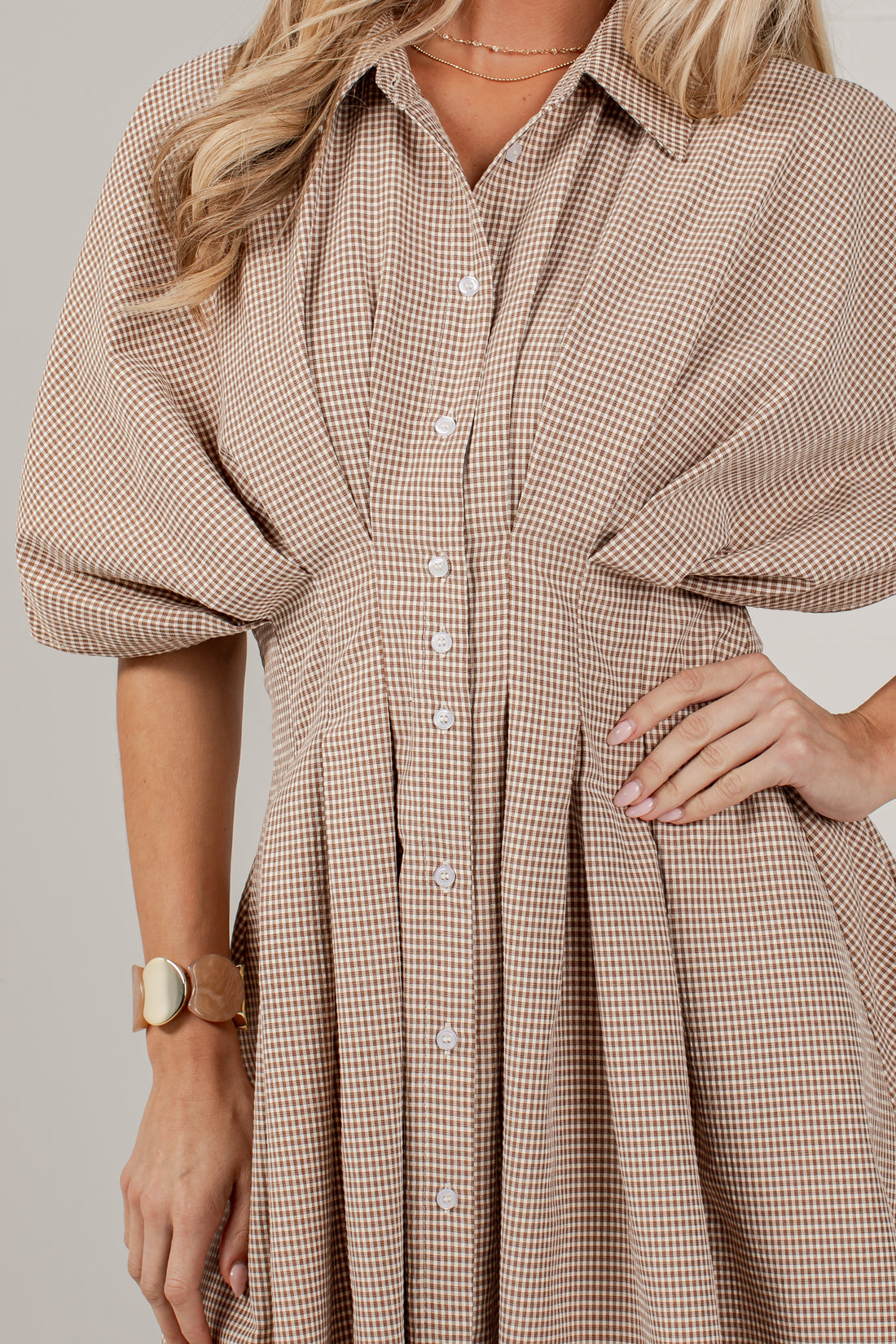 A close-up of a woman wearing a beige and brown gingham button-up shirt dress with voluminous elbow-length sleeves and a pleated skirt, photographed against a plain light gray studio background.