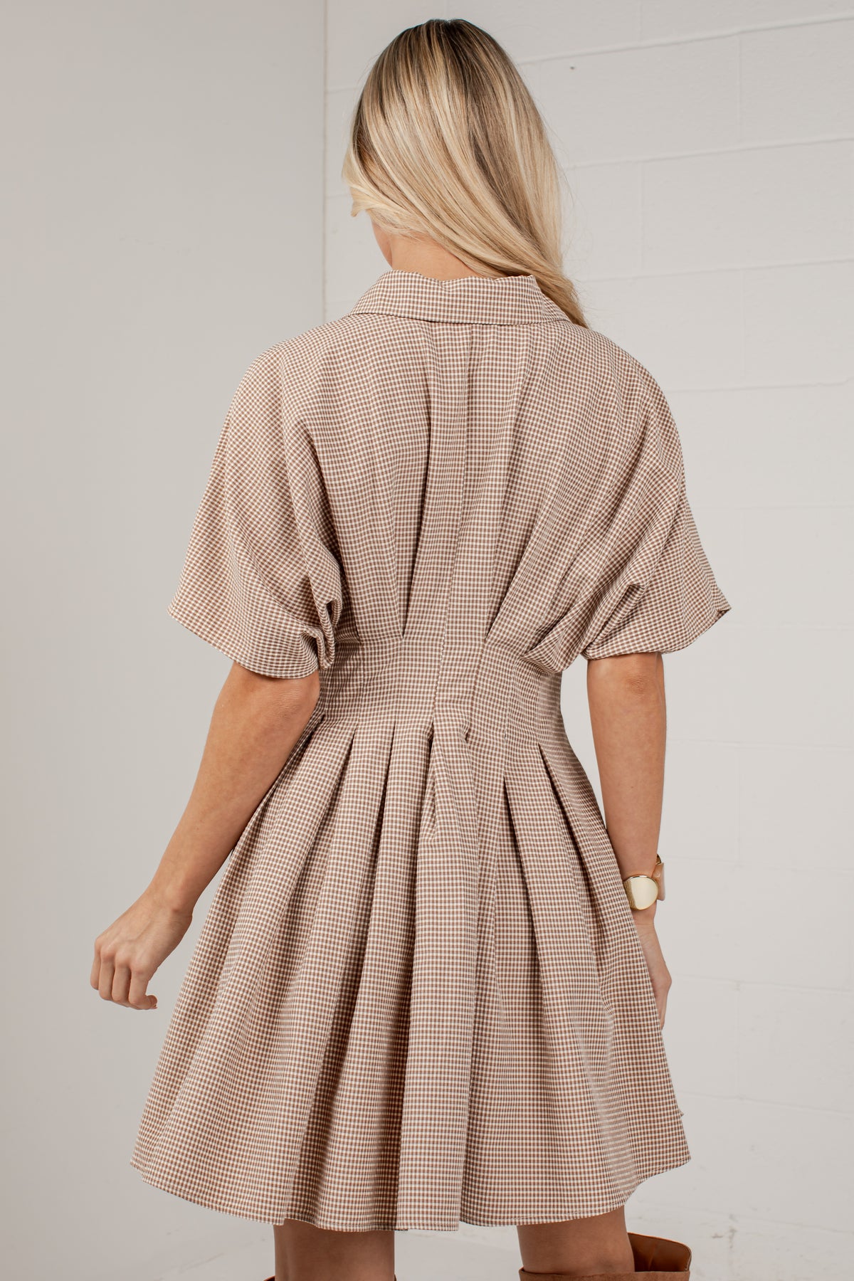 A woman shown from the back wearing a beige and brown checkered short-sleeve dress with a pleated skirt, set against a white paneled wall.