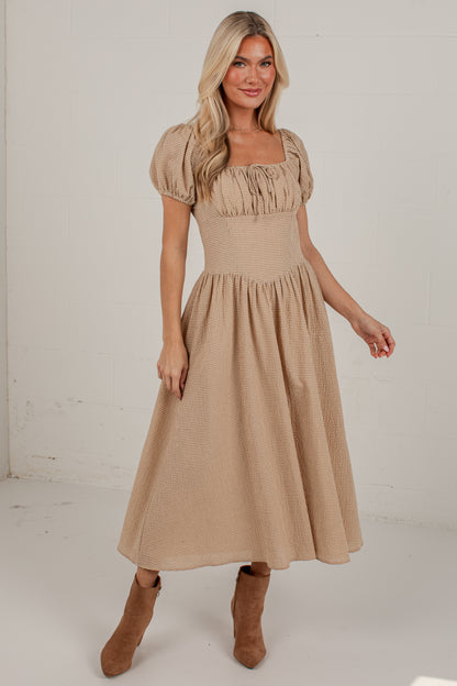 A woman modeling a beige midi dress with puff sleeves, a square neckline with gathered ruffles, and a flared skirt, paired with brown suede ankle boots against a white studio background.