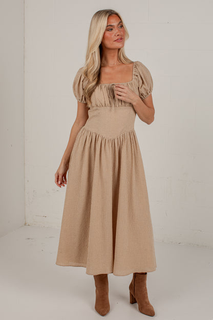 A woman modeling a beige midi dress with puff sleeves, a square neckline, and a gathered skirt, paired with brown ankle boots in a bright studio setting.