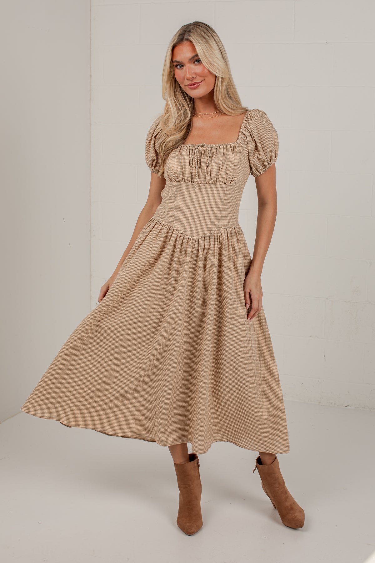 A woman modeling a beige midi-length dress with puff sleeves, a square neckline, and a gathered skirt, paired with brown ankle boots, set against a white studio background.