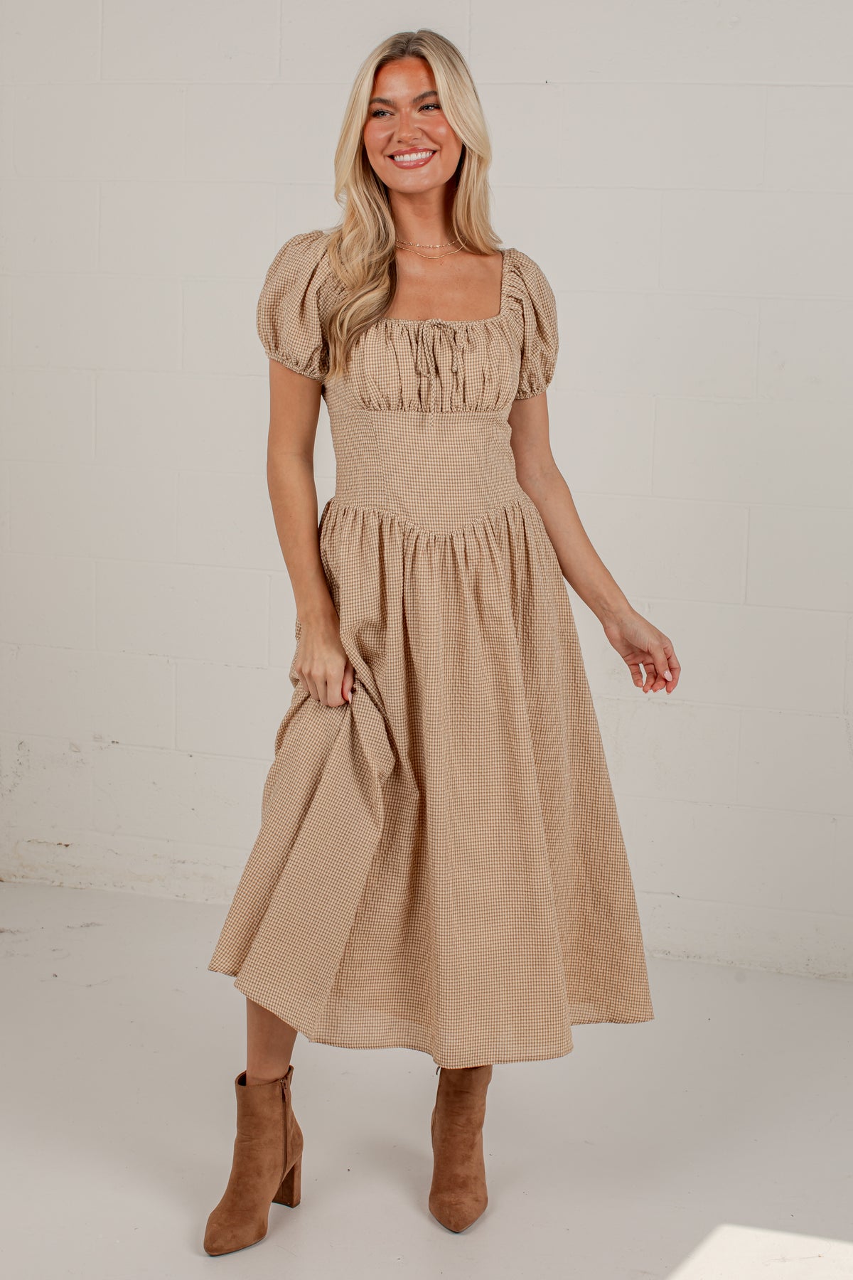 A woman modeling a beige midi dress with puff sleeves, a square neckline, and a gathered skirt, paired with brown ankle boots against a white studio background.