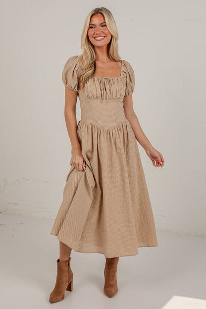 A woman modeling a beige midi dress with puff sleeves, a square neckline, and a gathered skirt, paired with brown ankle boots against a white studio background.