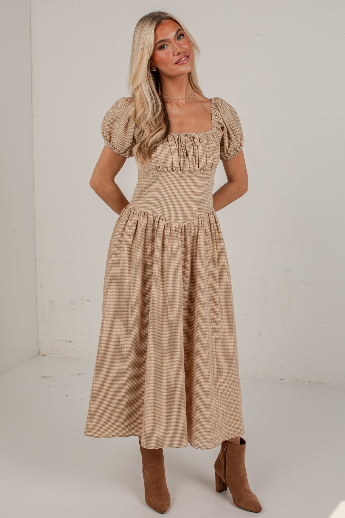 A woman modeling a beige midi dress with puff sleeves, a square neckline, and a gathered skirt, paired with brown ankle boots against a white studio background.