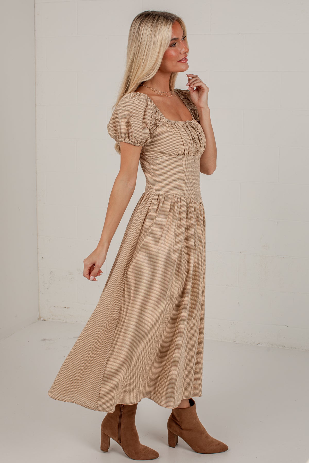 A woman modeling a beige midi-length puff-sleeve dress with a gathered bodice and flared skirt, paired with brown suede ankle boots, set against a white brick wall background.