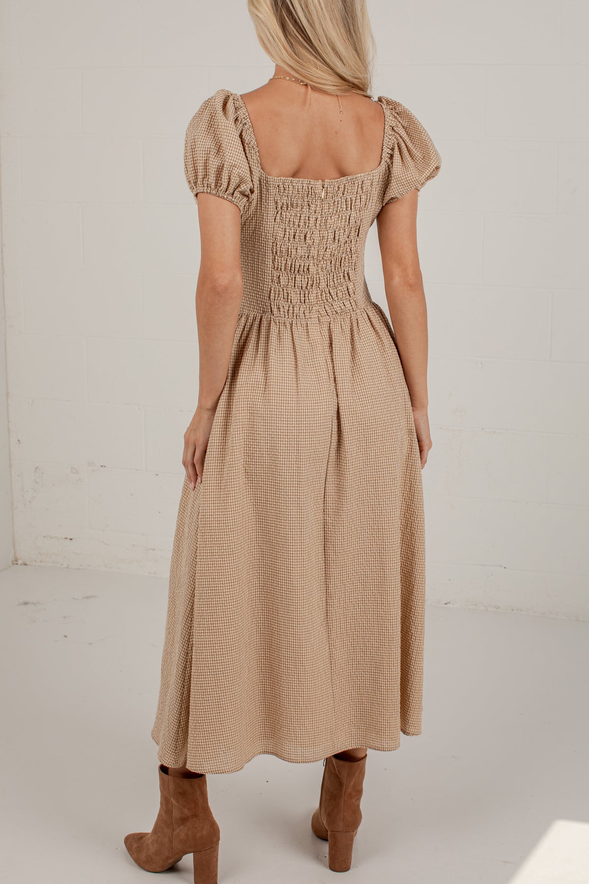 A woman viewed from the back wearing a beige midi dress with puff sleeves, a smocked bodice, and a gathered skirt, paired with brown ankle boots in a bright studio setting.
