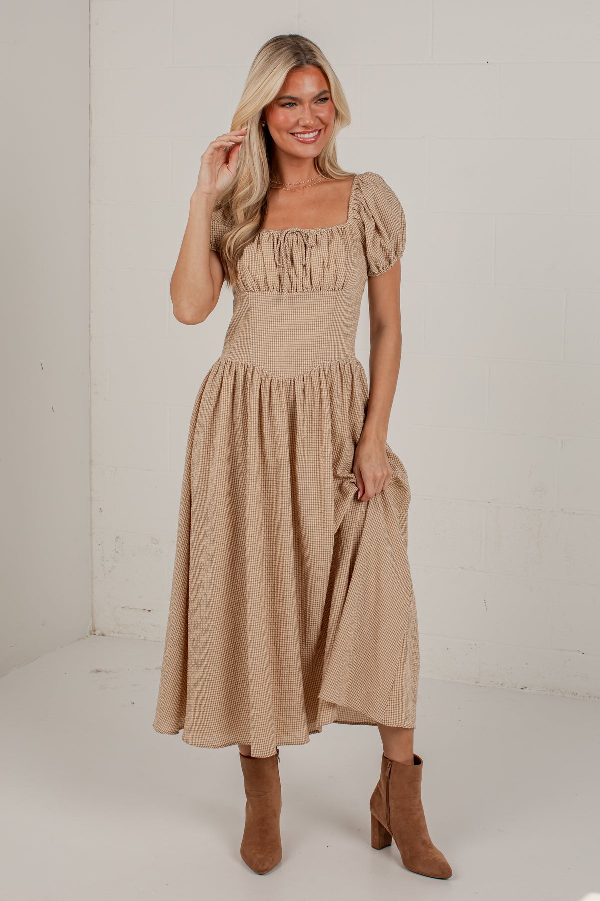 A woman modeling a beige midi dress with puff sleeves, a square neckline, and a gathered skirt, paired with brown ankle boots in a bright studio setting.
