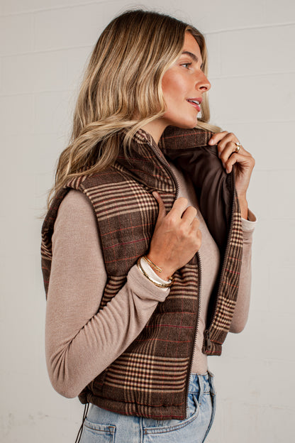 A woman wearing a brown plaid zippered vest over a beige long-sleeve top and light blue jeans, standing against a white paneled wall.