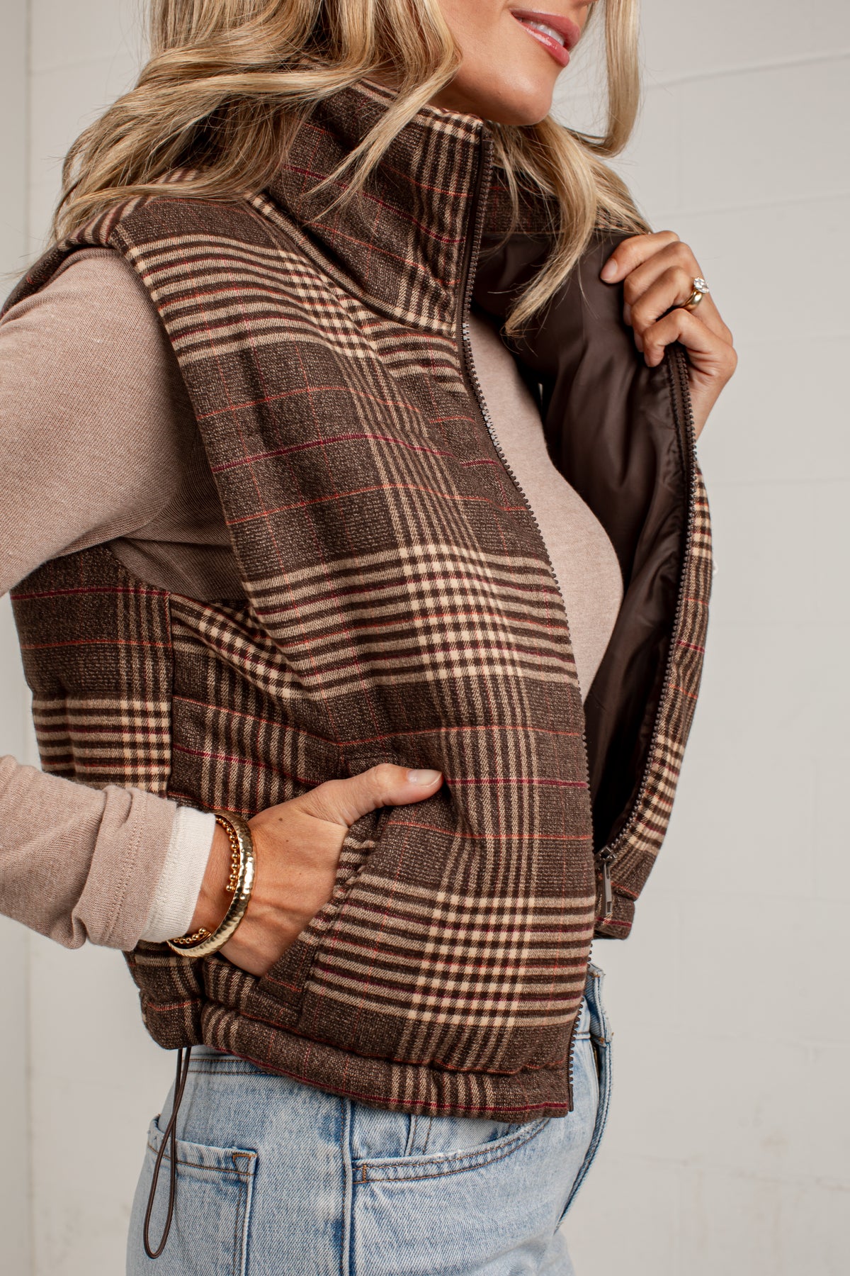 A woman wearing a brown plaid zip-front vest over a beige long-sleeve top and light blue jeans, shown in a close-up lifestyle shot against a white wall.