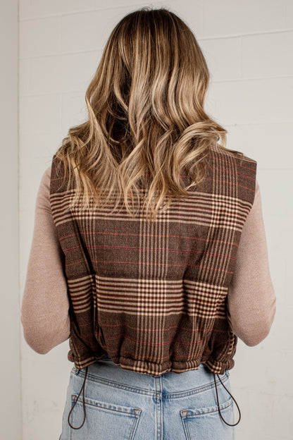 A woman viewed from behind wearing a brown plaid sleeveless vest over a beige long-sleeve top and light blue jeans, standing against a white paneled wall.