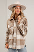 A woman wearing a beige wide-brim hat and a brown and cream plaid flannel button-up shirt jacket, paired with light blue jeans, standing against a white brick wall.