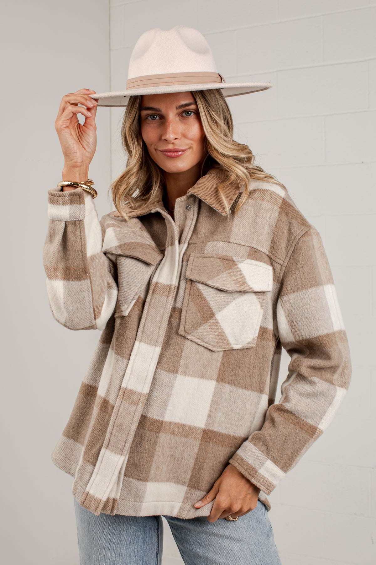 A woman wearing a beige and brown plaid flannel button-up shirt jacket with a wide-brimmed cream hat, posing against a light gray brick wall.