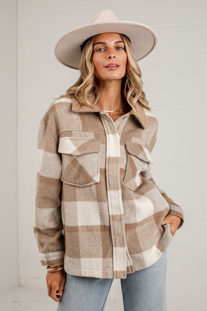 A woman wearing a beige and white buffalo plaid button-up shirt jacket, a wide-brimmed beige hat, and light blue jeans, standing against a white paneled wall.