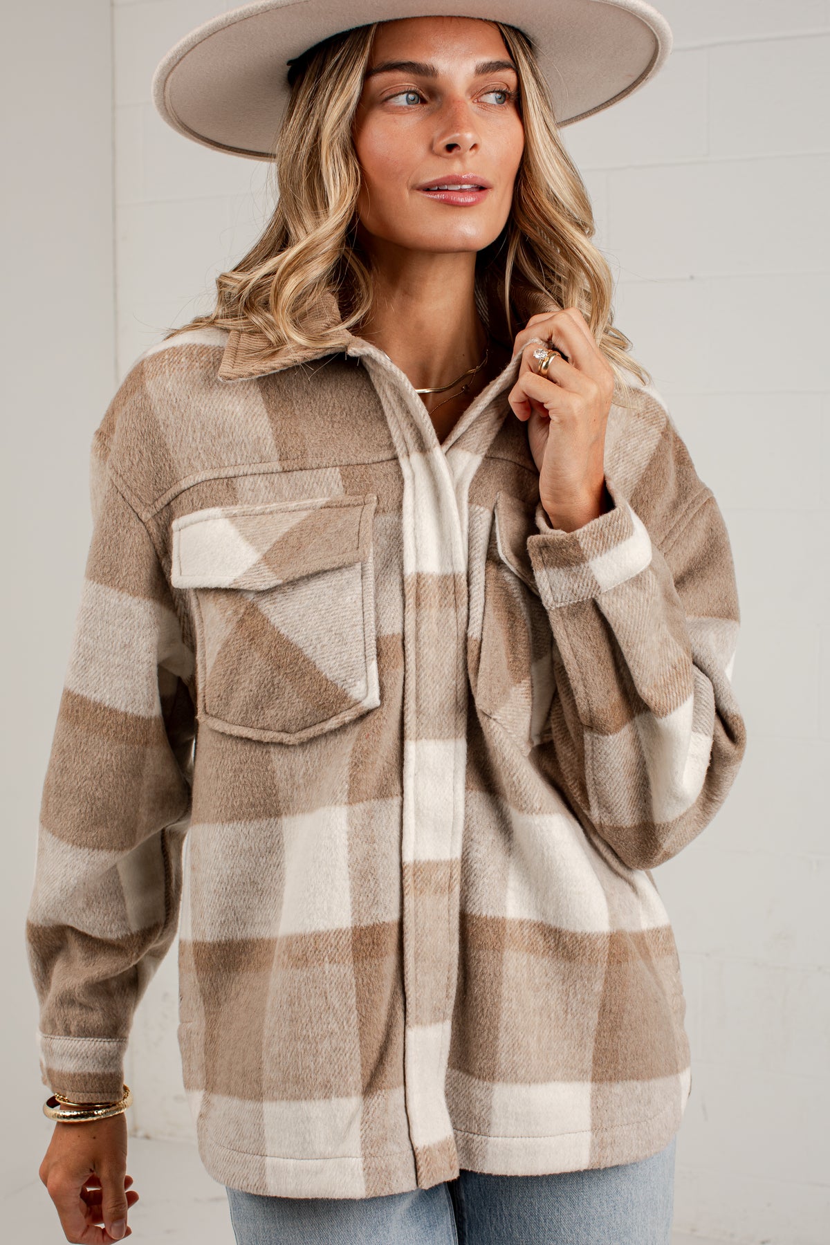 A woman wearing a beige and cream plaid button-up shirt jacket with a wide-brim hat, standing against a white paneled wall.