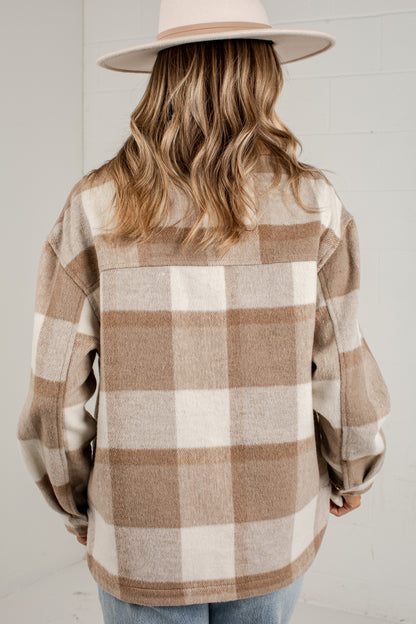A woman viewed from behind wearing a beige and off-white buffalo plaid flannel shirt jacket, a light beige wide-brim hat, and blue jeans against a white paneled wall.