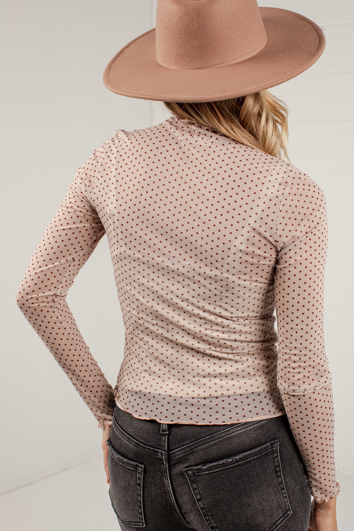 A woman shown from the back wearing a fitted long-sleeve top with a small repeating dot pattern in a warm beige/taupe tone, paired with dark gray jeans and a wide-brim tan hat against a plain light background.