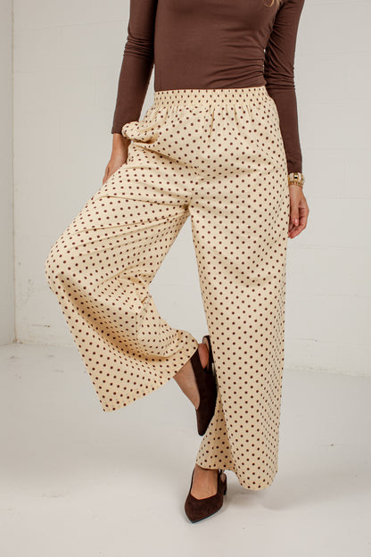 A person wearing high-waisted, wide-leg pants with a beige base and small brown polka dots, paired with a brown long-sleeve top and dark brown pointed flats, shown against a plain white studio background.