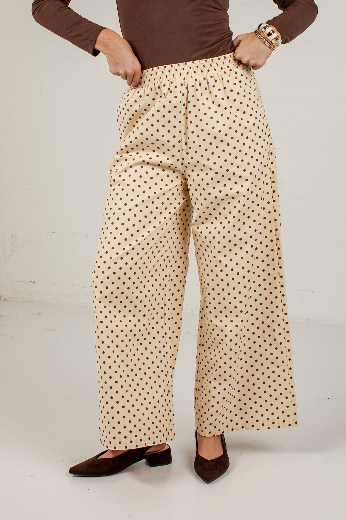 A person wearing high-waisted, wide-leg trousers with a beige base and small black polka dots, styled with a brown long-sleeve top and dark brown pointed-toe flats against a white studio background.