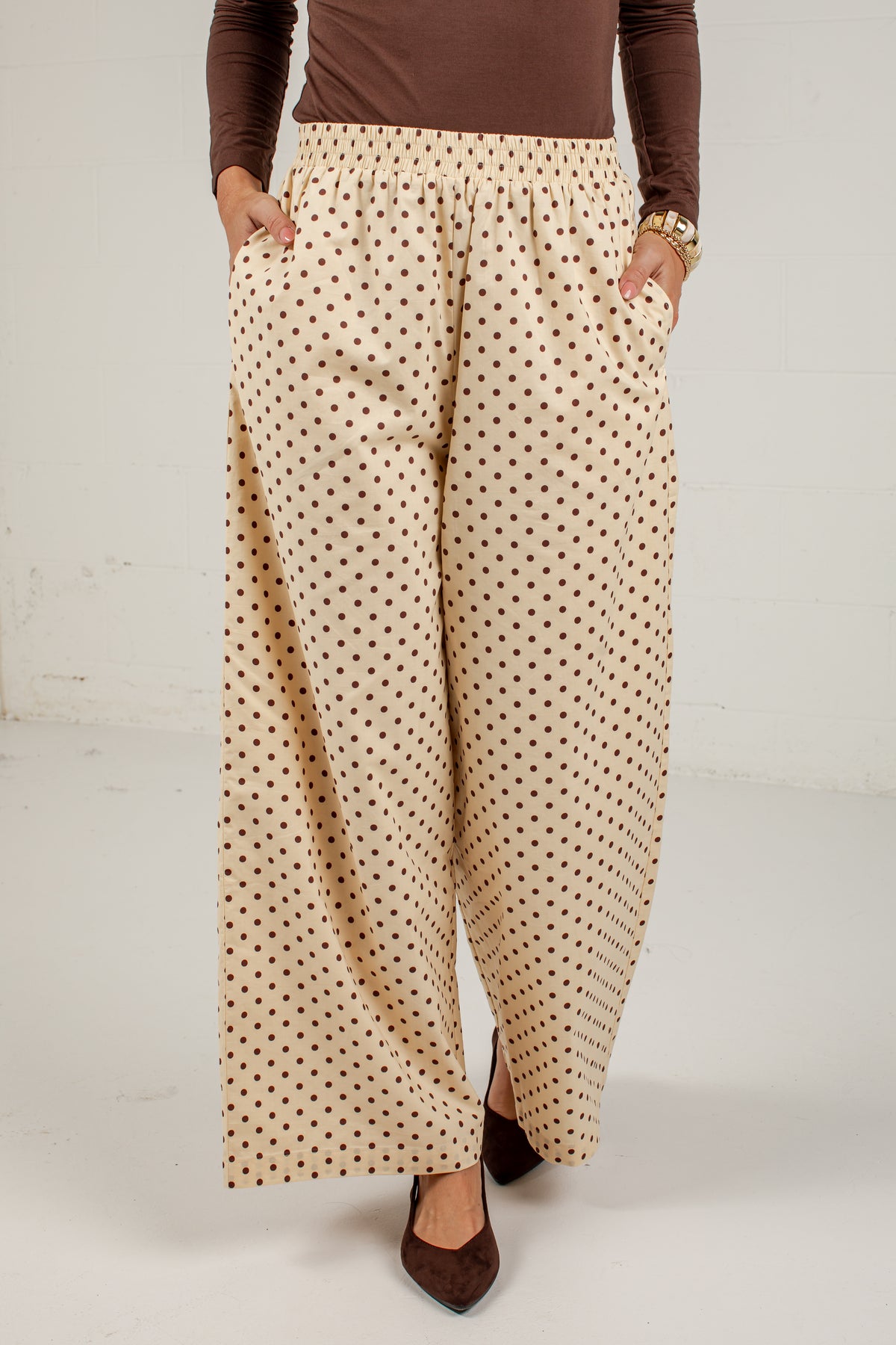 A person wearing wide-leg pants with a beige base and small brown polka dots, paired with a brown long-sleeve top and brown pointed shoes, standing against a plain light wall.