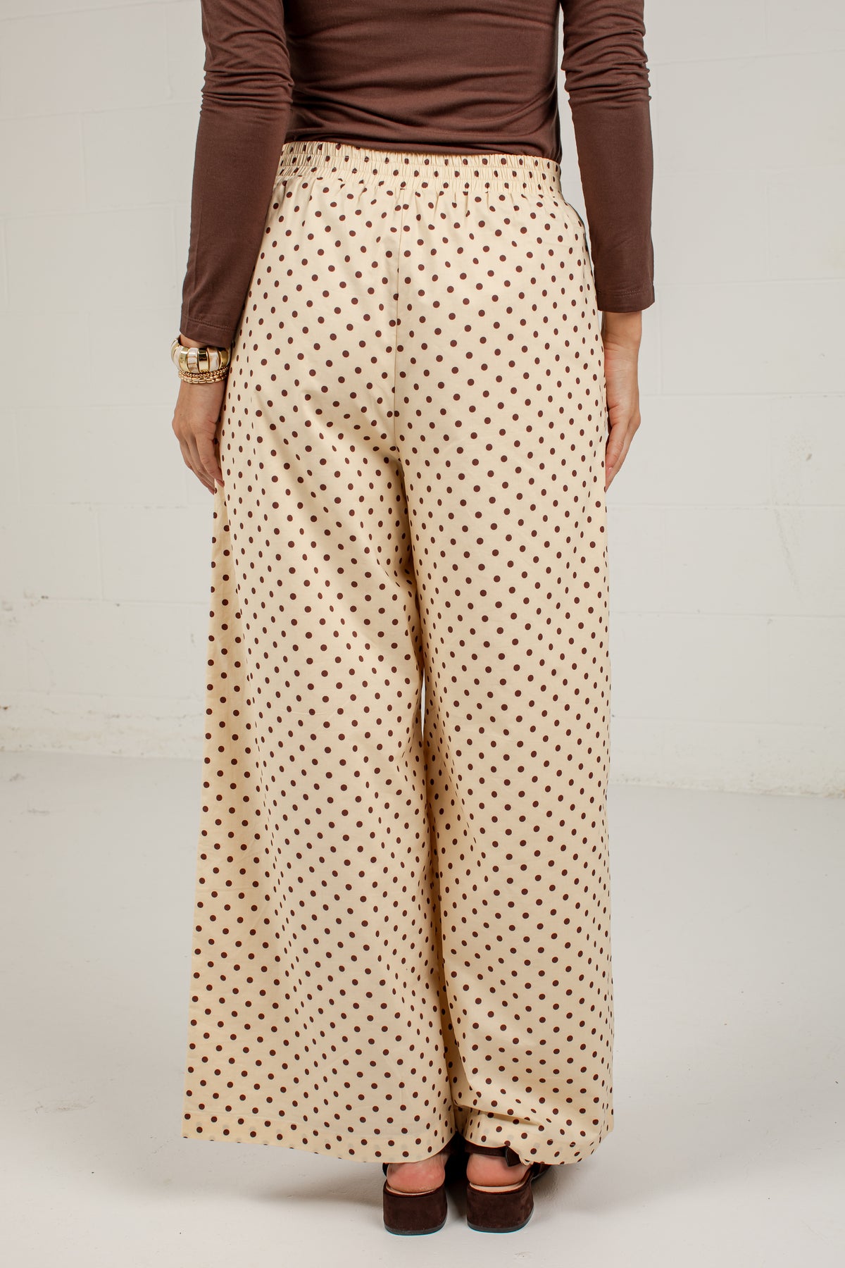 A person shown from the back wearing wide-leg cream pants with a small brown polka dot pattern, a brown long-sleeve top, and brown platform sandals against a plain light gray studio background.
