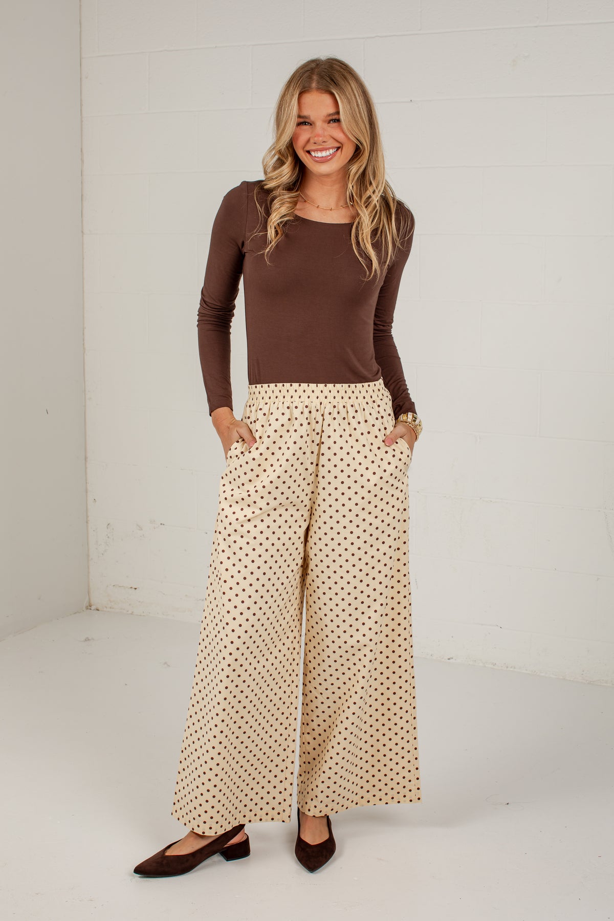 A woman modeling a brown long-sleeve top and beige wide-leg pants with a small black polka dot pattern, standing against a white wall.