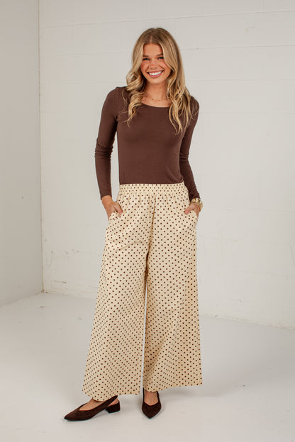 A woman modeling a brown long-sleeve top and beige wide-leg pants with a small black polka dot pattern, standing against a white wall.