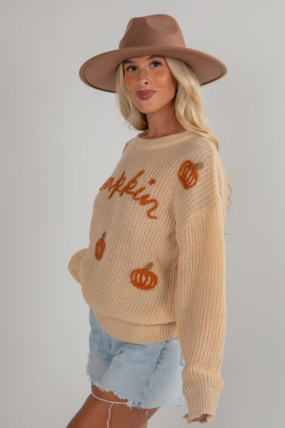 Woman wearing a beige sweater with pumpkin graphics and a brown hat on a plain background